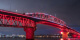 Image of the Auckland Harbour Bridge with pink and red lights at night, with the Auckland cityscape in the background