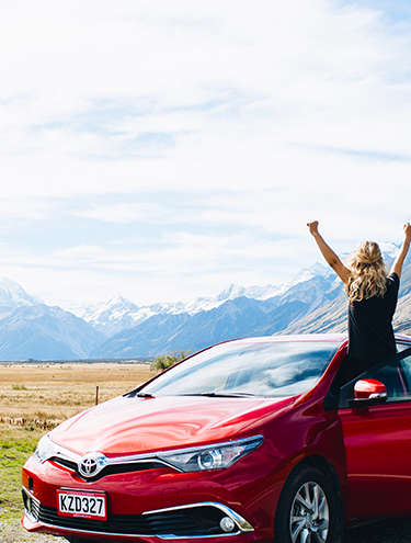 Image featuring a woman standing from the side of the front seat of her GO Rentals car with her arms in the air in excitement, looking out to a scenic Queenstown snow-capped mountain backdrop