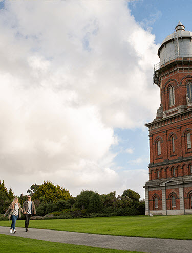 Two people walking along a path next to the historic Invercargill Water Tower, a tall red-brick structure with a domed top, set against a clear blue sky with scattered clouds.