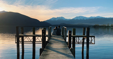 A couple sitting on a dock at sunset, overlooking a calm lake with snow-capped mountains in the background in Te Anau.