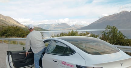 Person entering a white GO Rentals car by a lakeside with mountains in the background