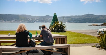 Two people sitting at a picnic table by the water, with hills and boats in the distance