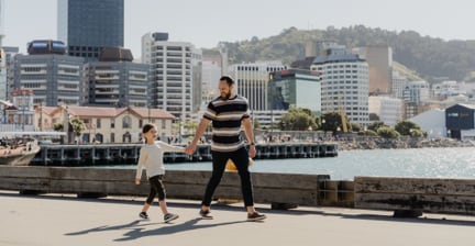 Father and child walking on a waterfront with city buildings in the background