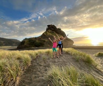 Three people standing on a sandy hill, celebrating at the beach during sunset