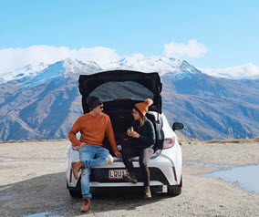 Two people sitting in the back of a white car with mountains in the background