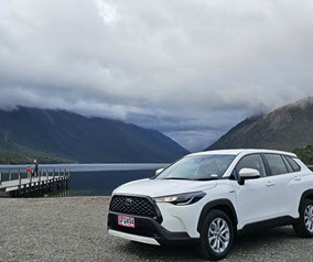 White SUV parked near a lake with mountains and a dock in the background