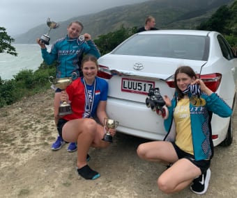 Three people posing with trophies next to a white car on a cliffside road