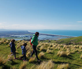 Person walking with two children on a grassy hill overlooking the ocean