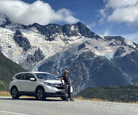 Person standing by a white car with a backdrop of snow-covered mountains